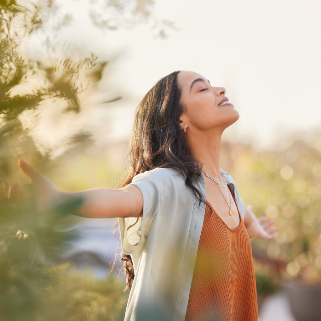 Woman joyfully extends arms, eyes closed, enjoying sunlight in a lush garden.