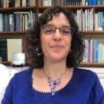 Person with curly hair and glasses, wearing a blue top, sitting in front of a bookshelf.