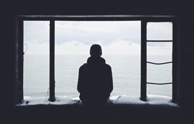 Person sitting in a window frame, looking out at a calm sea under an overcast sky.