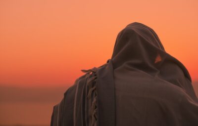 Silhouetted person draped in cloth gazing at a vibrant orange sunset.