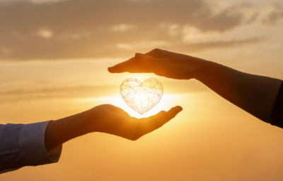 Two hands hold a heart-shaped wire object against a sunset sky, symbolizing love and connection.