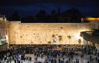 Crowd gathering at the Western Wall during the evening, with a lit stone wall and night sky in the background.
