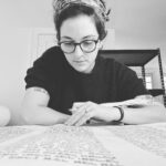 Woman with glasses reading a large document on a table, black and white photo.