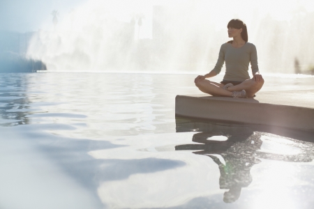 A person sitting cross-legged by the edge of a calm pool with mist in the background.