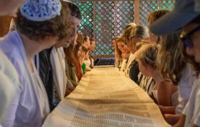 People gathered around a long table, reading a scroll in a wooden pavilion.