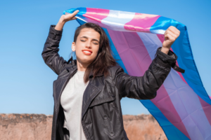 Person joyfully holding a transgender pride flag against a clear blue sky background.