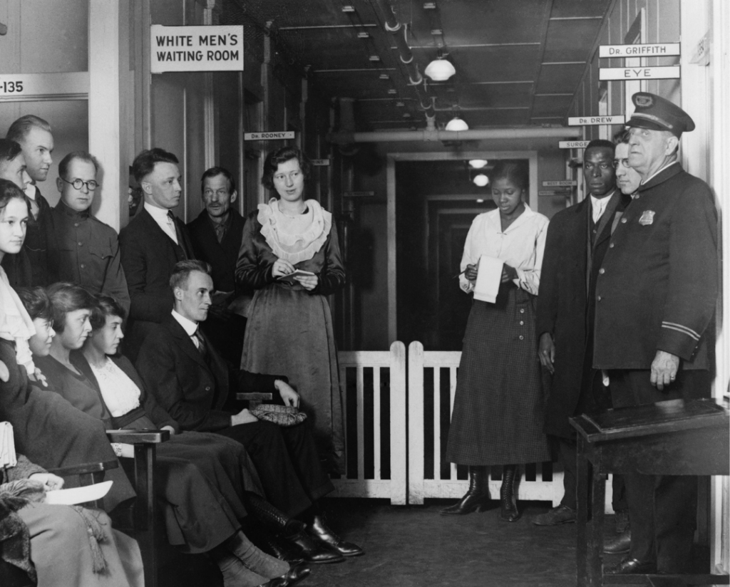 A segregated waiting room with a White Mens Waiting Room sign, showing people seated and some standing, including a guard.
