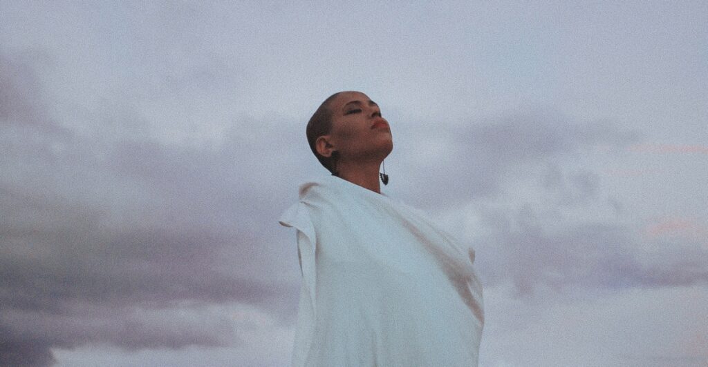 Person in a white outfit stands against a cloudy sky, looking upwards with a serene expression.
