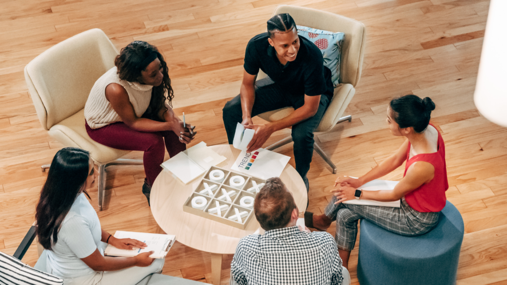 Five people having a meeting, sitting in a circle with notepads and coffee cups, engaged in discussion.