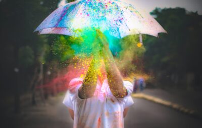 Person under umbrella with colorful powder bursting around, standing outdoors on a blurred street.