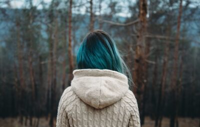 Person with teal hair in a beige sweater stands facing a forest of tall, leafless trees.