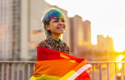 Person with colorful hair smiles, wrapped in a rainbow flag, standing outdoors during sunset.