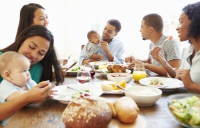 A diverse group of people enjoying a meal together, with two adults holding babies at a dining table.