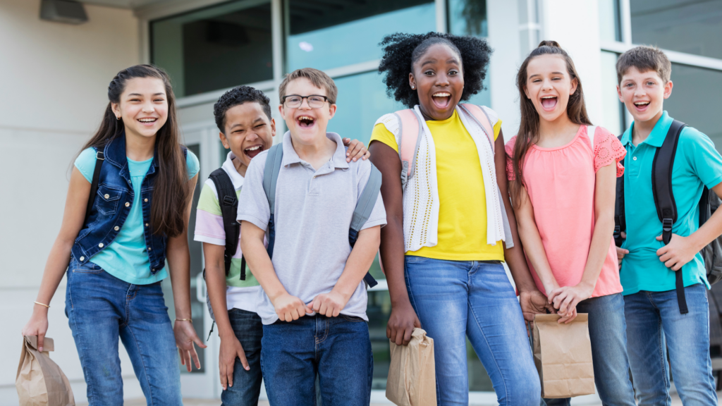 A group of joyful children with backpacks and lunch bags stand outside a building, smiling and laughing.