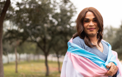 Smiling person wrapped in a transgender pride flag, standing in a park with trees in the background.