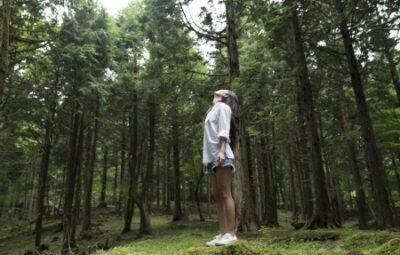A person stands in a forest, looking up at tall trees with a peaceful, contemplative expression.