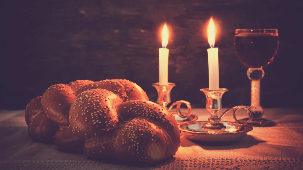 Challah bread, two lit candles, and a glass of wine on a table with a dark background.