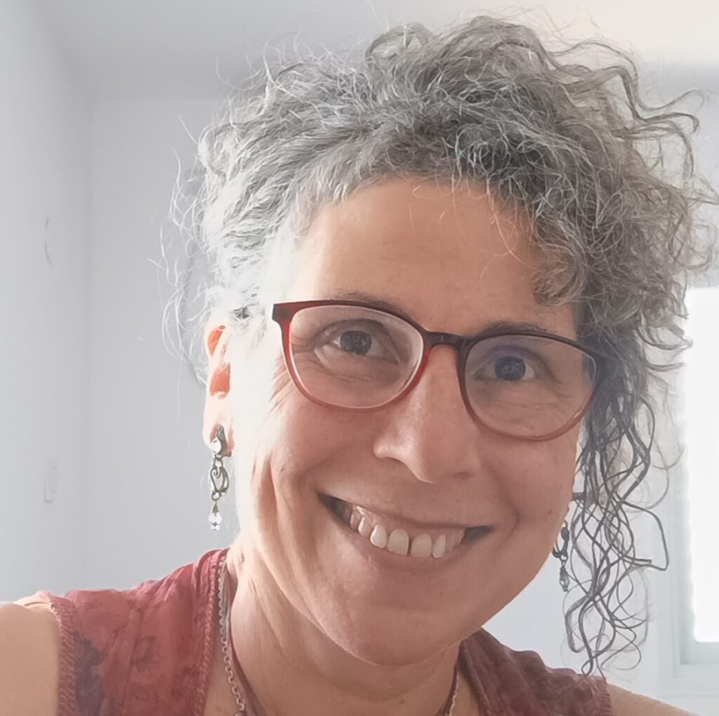 Smiling person with curly gray hair and red glasses, wearing earrings and a sleeveless top, in a bright room.
