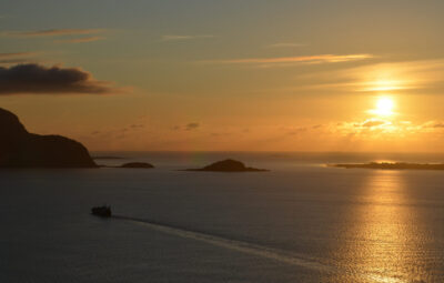 Sunset over calm sea with islands; a silhouette of a ship sails on the horizon under an orange and gold sky.