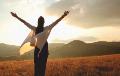 A person with outstretched arms stands in a field at sunset, embracing the natural landscape.