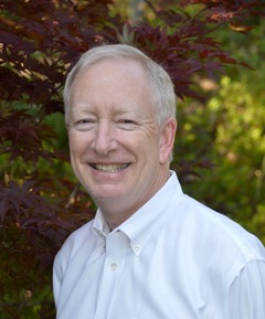 Smiling person with white hair wearing a white shirt, standing outside with leafy background.