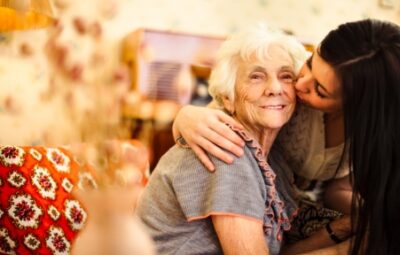 A young woman kisses an elderly woman on the cheek, both smiling warmly in a cozy room.