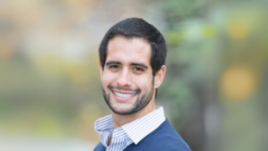 Justin Rosen Smolen smiles in front of a blurred outdoor background. He's wearing a blue sweater and a dress shirt with the top button undone.