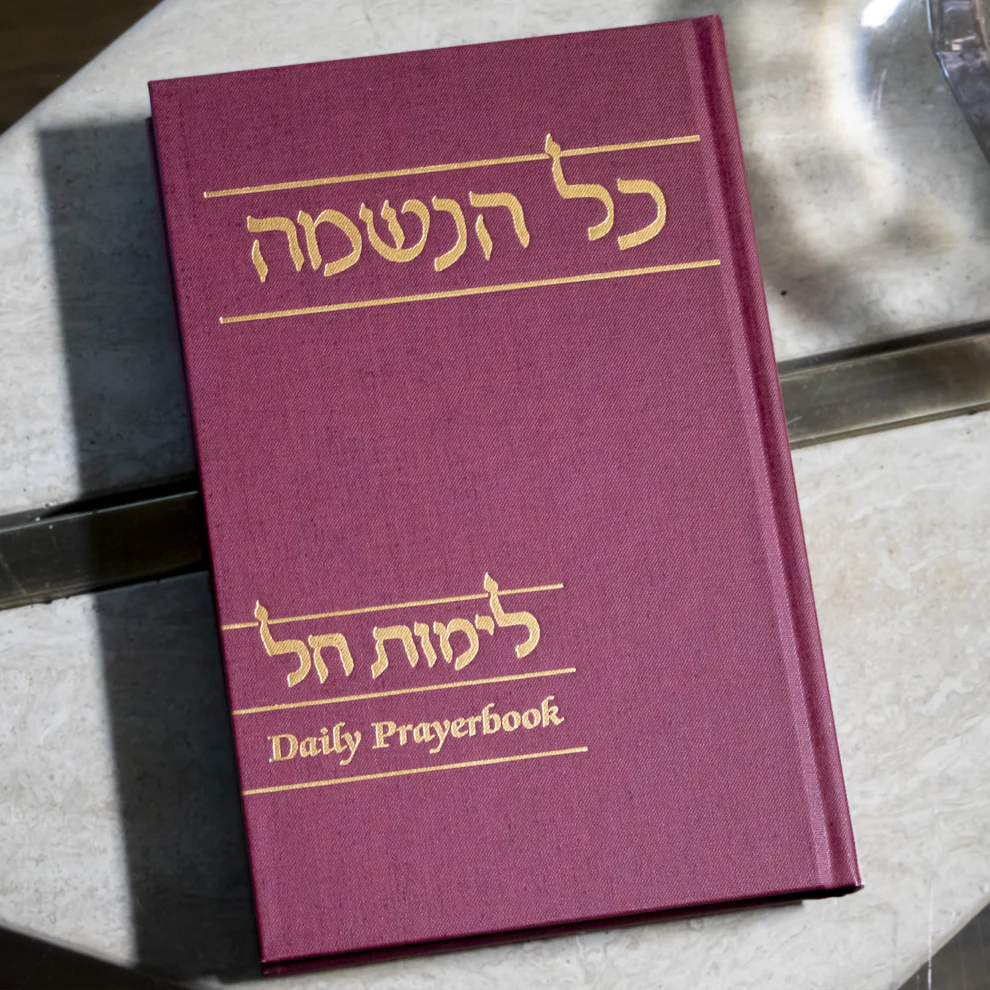 A maroon daily prayer book with gold embossed Hebrew and English text on a marble surface.