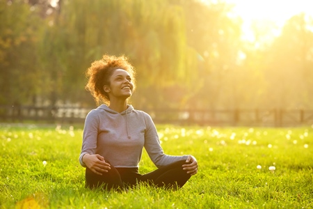 Person sitting cross-legged on grass in a park, smiling and looking up, with sunlight filtering through trees.