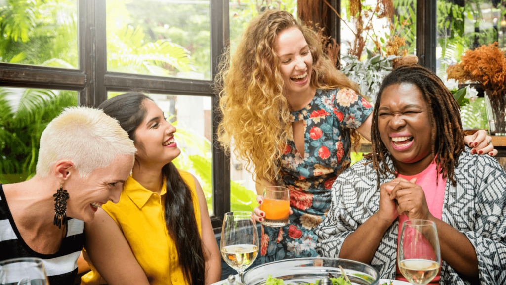 A group of women of different races laughing around a table with wine and dishes of food