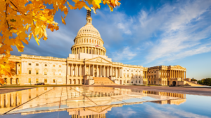 The U.S. Capitol building with golden autumn leaves and a reflective pool under a blue sky.