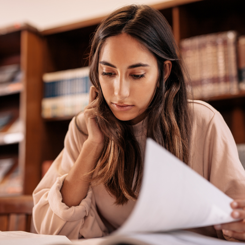 A woman reading documents at a table with bookshelves in the background.