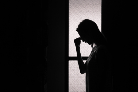 Silhouette of a person standing by a window, head bowed and hand on forehead, suggesting contemplation or stress.