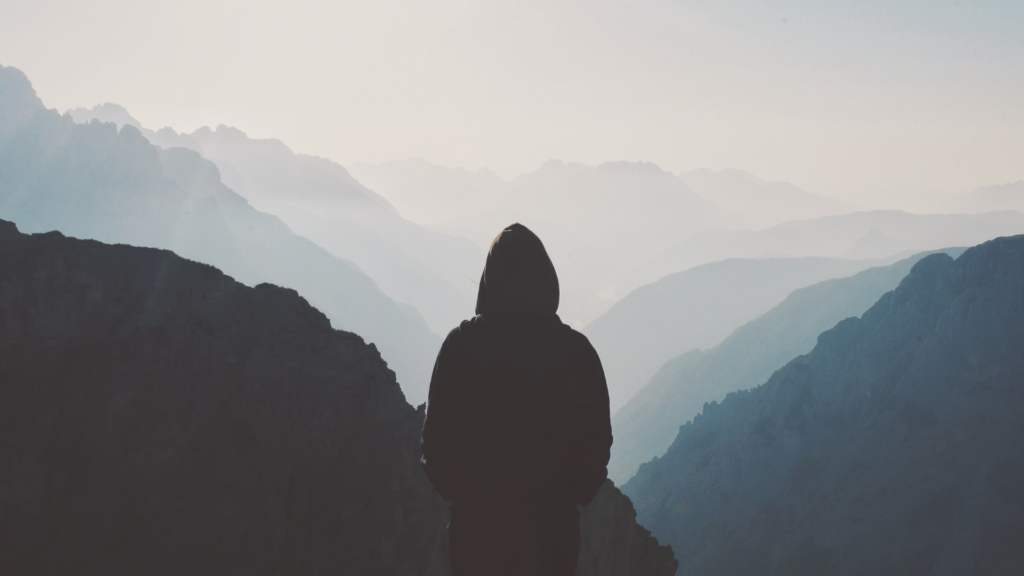 Silhouette of a person in a hoodie standing on a mountain, overlooking misty, layered mountain ranges.