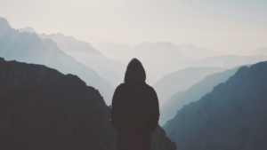 Silhouette of a person in a hoodie standing on a mountain, overlooking misty, layered mountain ranges.