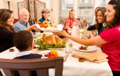 Family gathered around a table with a roasted turkey and various dishes, sharing a meal and smiling.