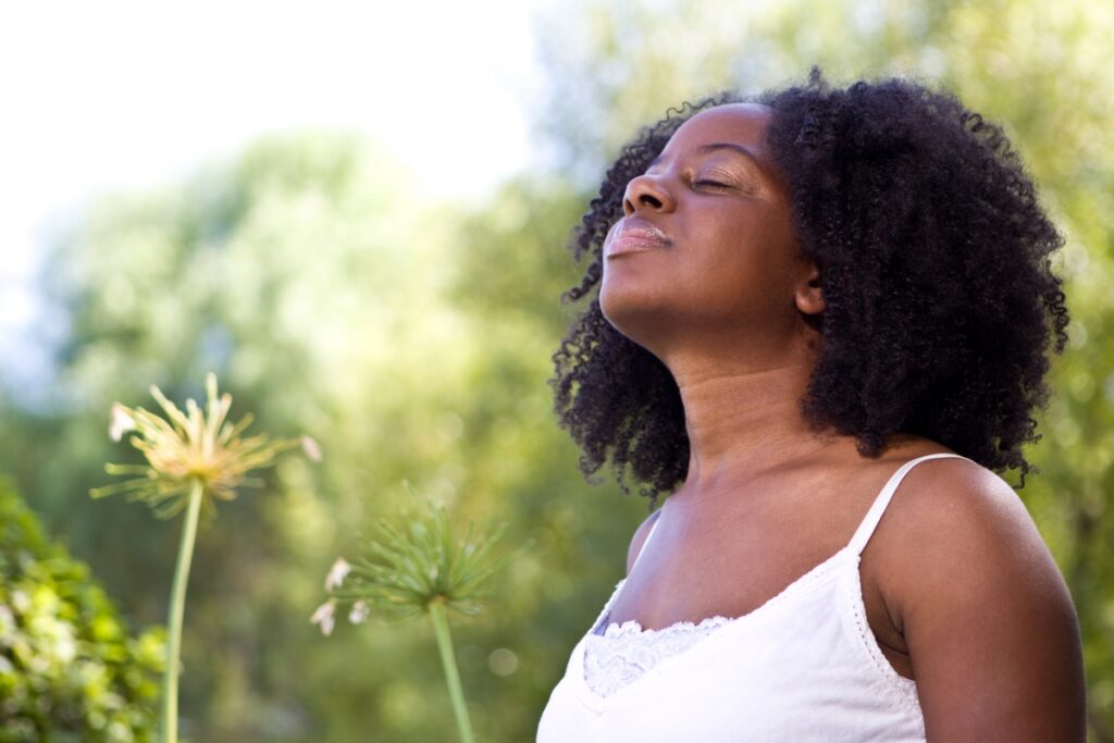 Woman in white top enjoying sunlight with eyes closed, surrounded by greenery.
