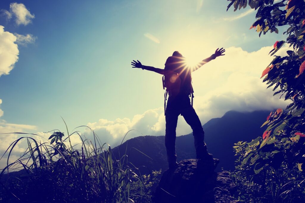Silhouetted hiker on a mountain top with arms outstretched, sun shining, surrounded by lush plants and cloudy sky.