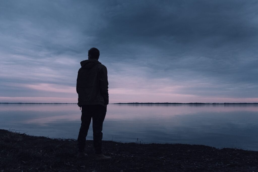 Silhouette of a person standing by a calm lake at dusk, with a cloudy sky above.