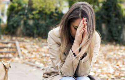 A woman sitting outside, hands covering her face, surrounded by fallen leaves.