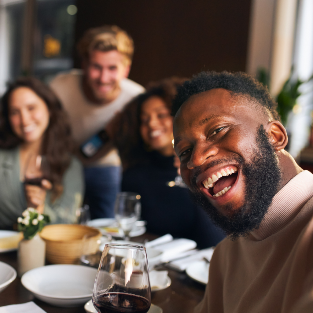 a black man, in focus at the front of the photo, sitting at a table with dinner on it and holding a glass while smiling. three other people, a white man and woman and a black woman, blurred in the background smiling behind the table.