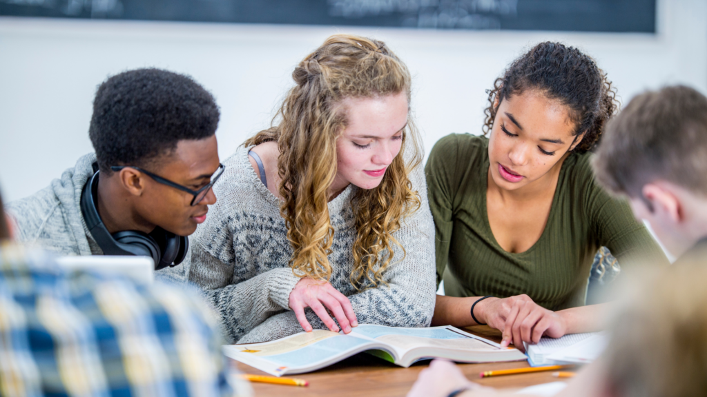 Three students studying together at a table, reading a textbook and taking notes.