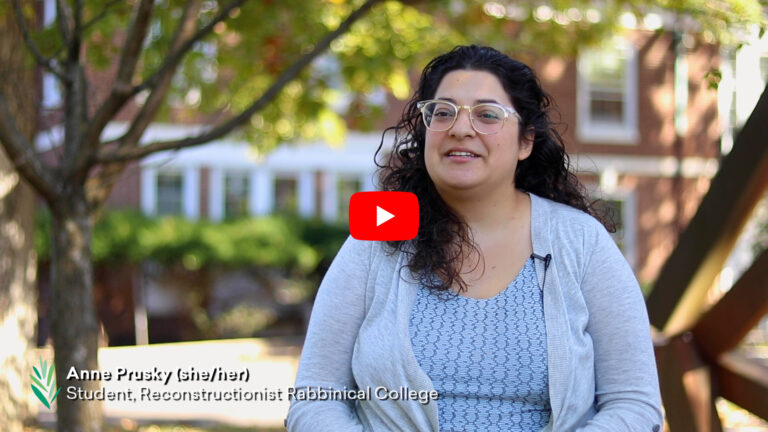 Anne Prousky, wearing glasses, sits outdoors, smiling, with trees and the RRC building in the background.