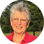 Smiling person with short gray hair and glasses, wearing a red shirt, stands outside in a park with trees.
