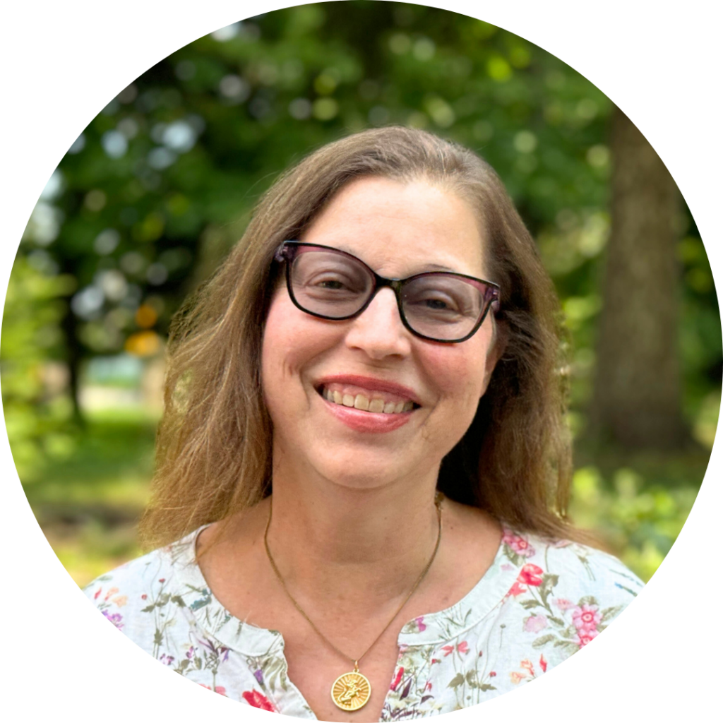 Smiling woman with glasses, wearing a floral shirt, stands outdoors with trees in the background.