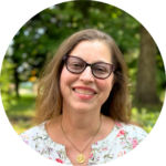 Smiling woman with glasses, wearing a floral shirt, stands outdoors with trees in the background.