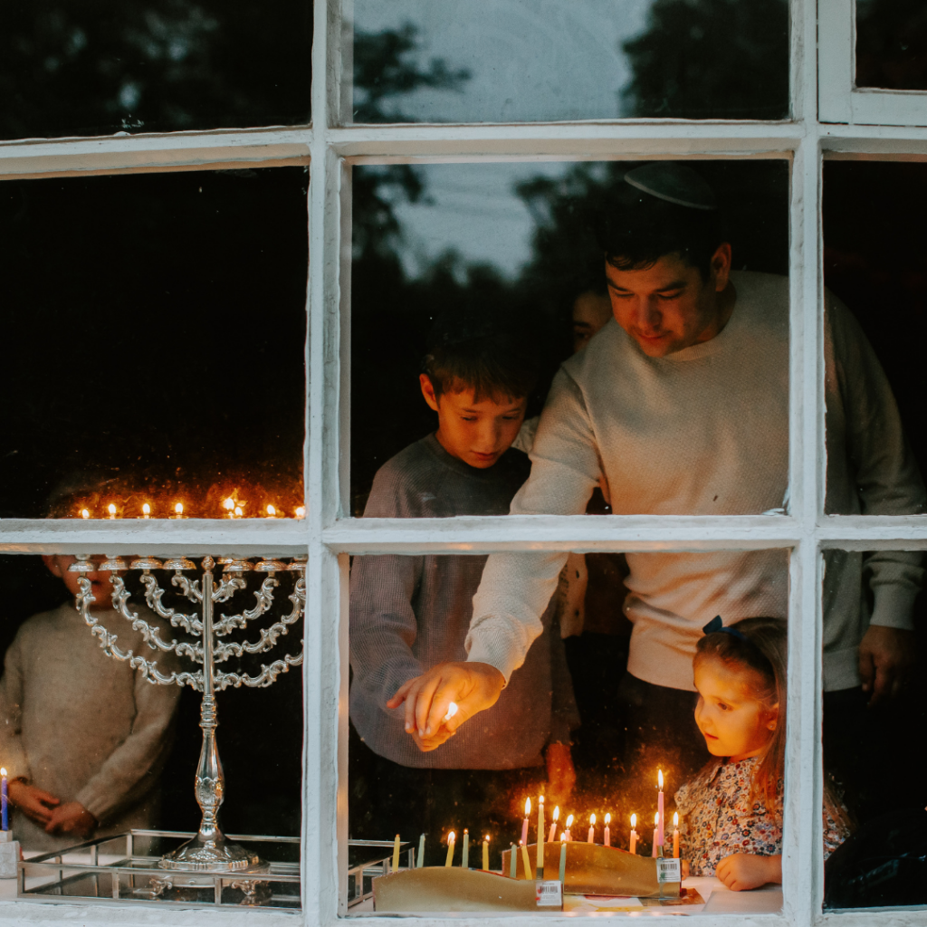 Family lighting menorah candles seen through a window, with reflection of lit menorah in the glass.