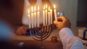 A person lighting the candles on a Hanukkah menorah, with coins and presents on the table.