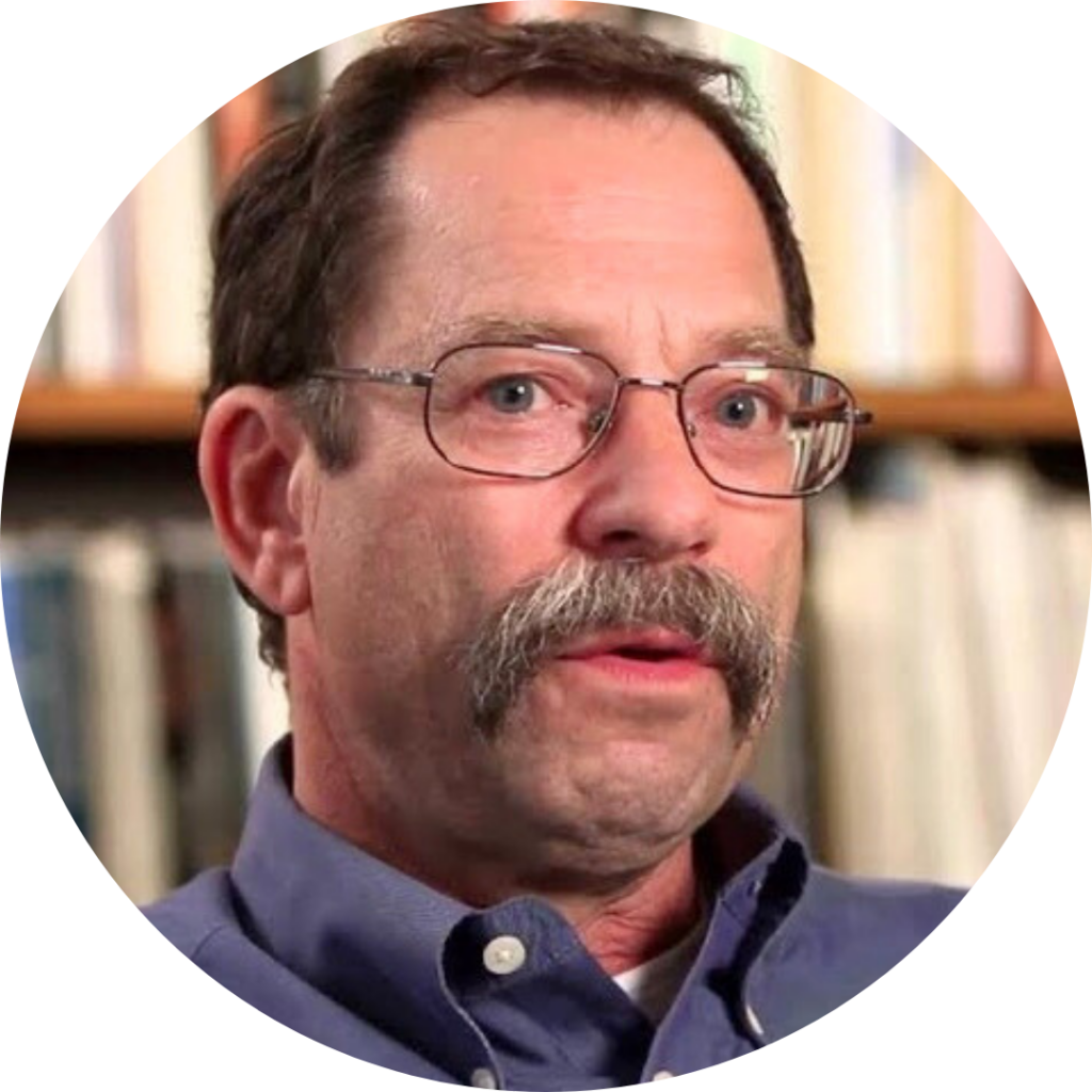 A man with glasses and a mustache in a blue shirt, sitting in front of a blurred bookshelf background.