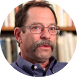 A man with glasses and a mustache in a blue shirt, sitting in front of a blurred bookshelf background.